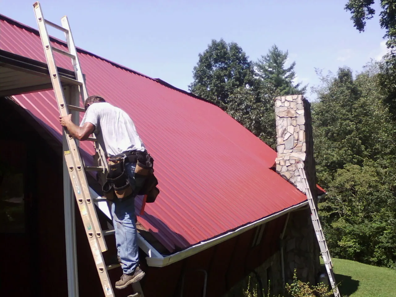 Roofer on a ladder beside a red metal roof and stone chimney — Soffit & Fascia Repair by Roofing Brothers in Nunda